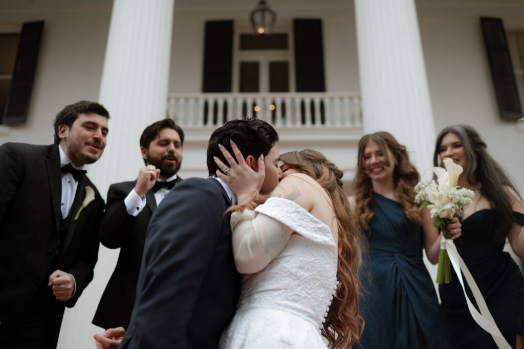 bride and groom kissing with the bridesmaids and groomsmen cheering in the background at Woodbine Mansion photo taken by Austin Wedding Photographer