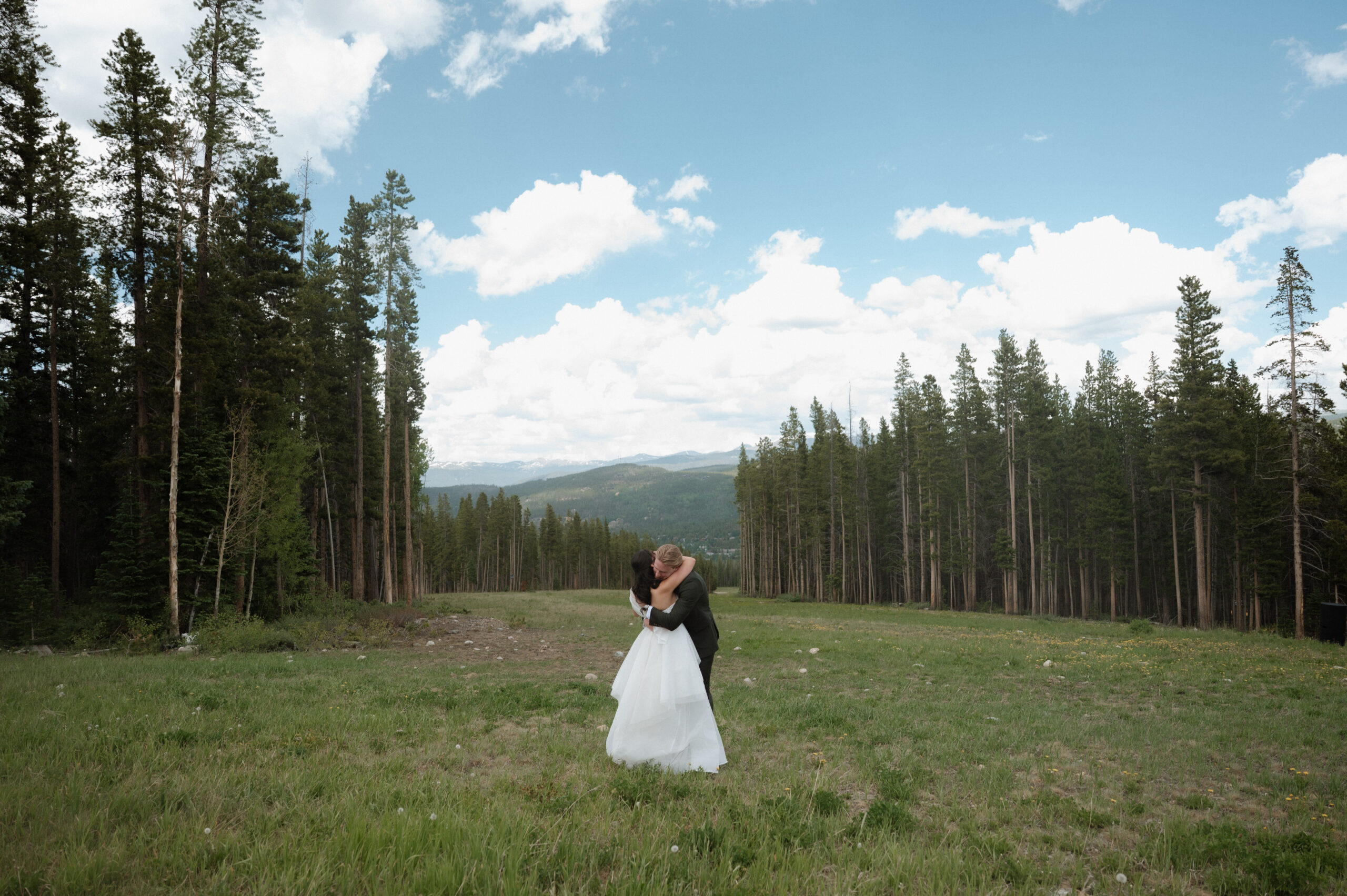 Bride and groom embrace in a wide-open field surrounded by pine trees and mountain views at their TenMile Station wedding in Breckenridge, Colorado.